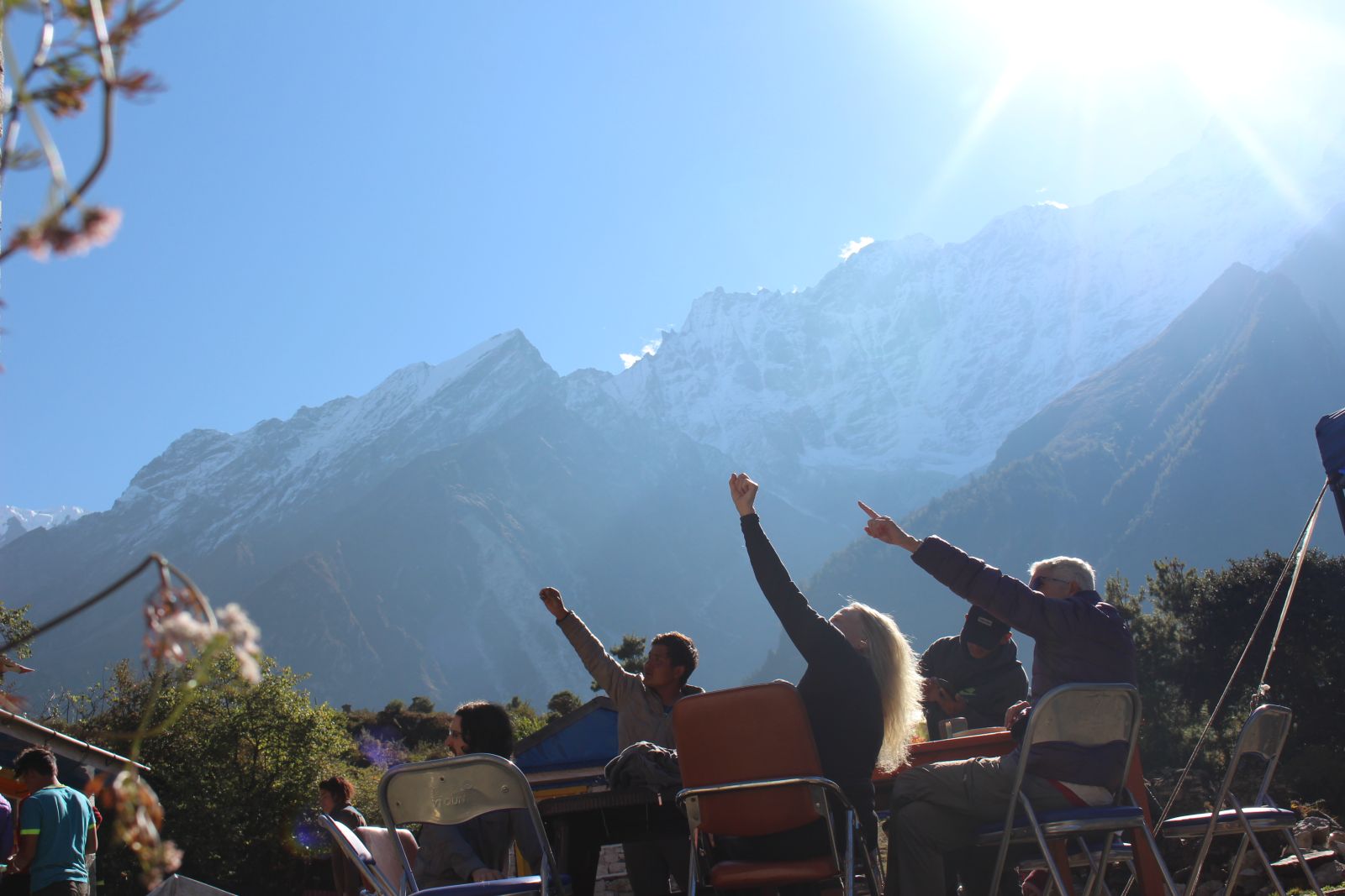 Few foreigner from west are sitting on a chair and table. There is a mountain backdrop and they are pointing to something to the top right left corner side of image. 