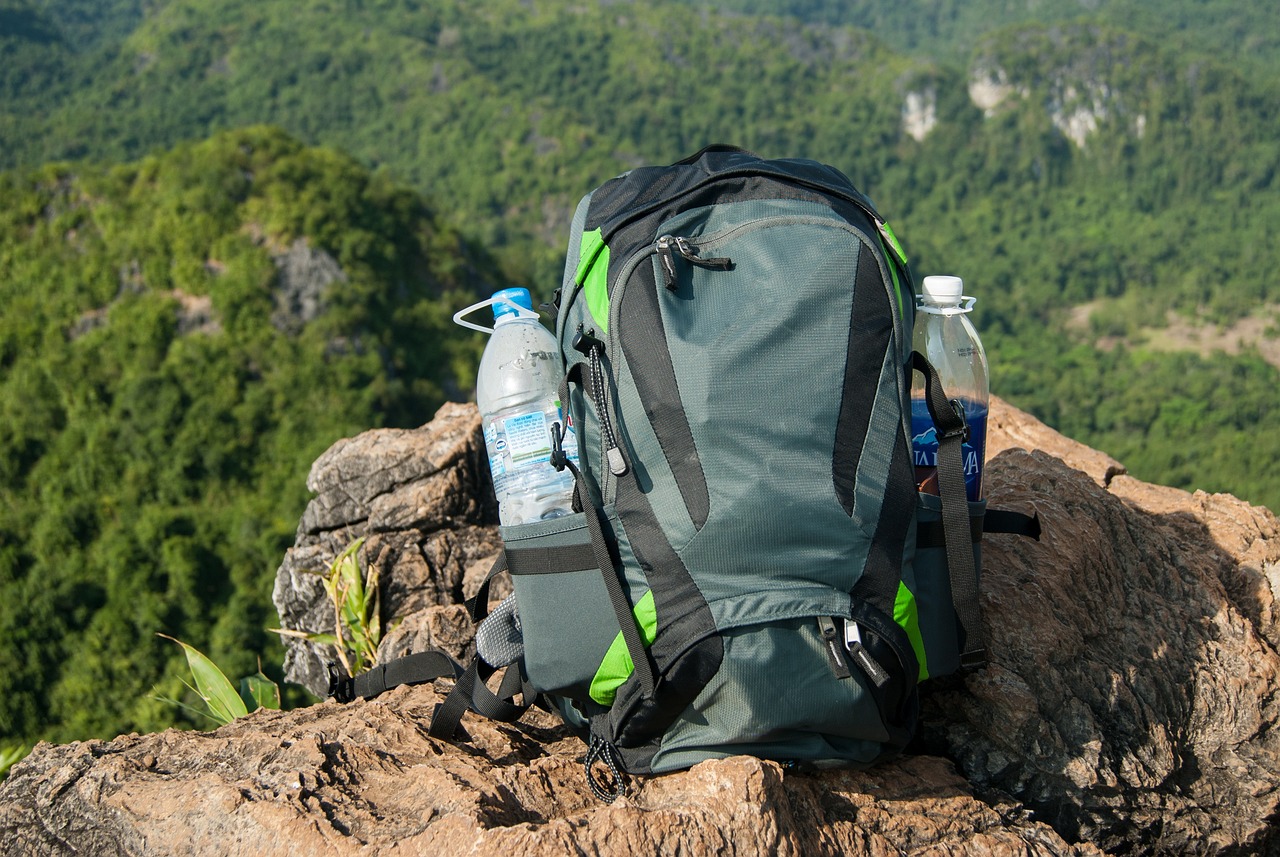 A gray hiking bag in which there is water bottle in both side pockets. It is placed at top of the hill on top of a giant rock. 