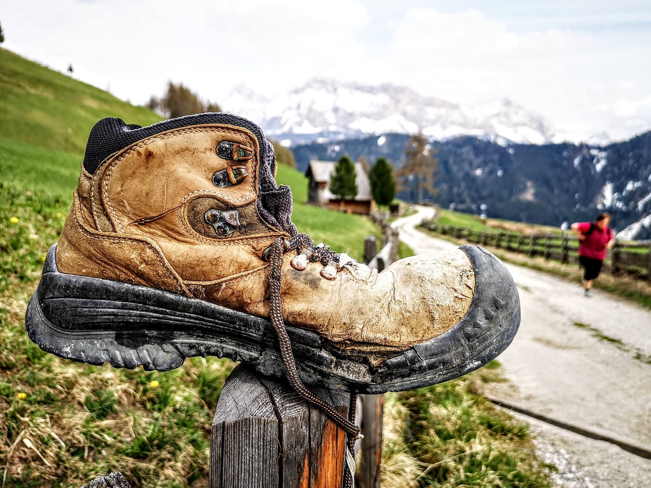 a dirty and brown trekking boot with black sole placed on a wooden fence. There is a blurred mountainous backdrop.