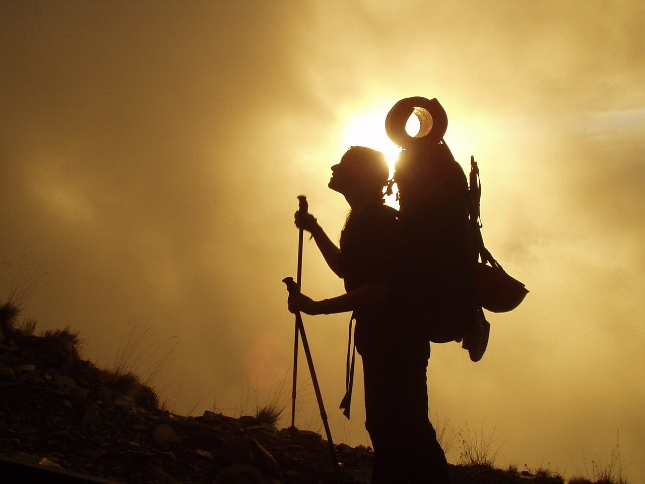 A man carrying a trekking bag with a pair of trekking pole looking upwards to the steep hill way ahead. The picture is in silhouette as the phot is clicked from the angle where camera and sun are in direct alignment and the man and hill in in between.