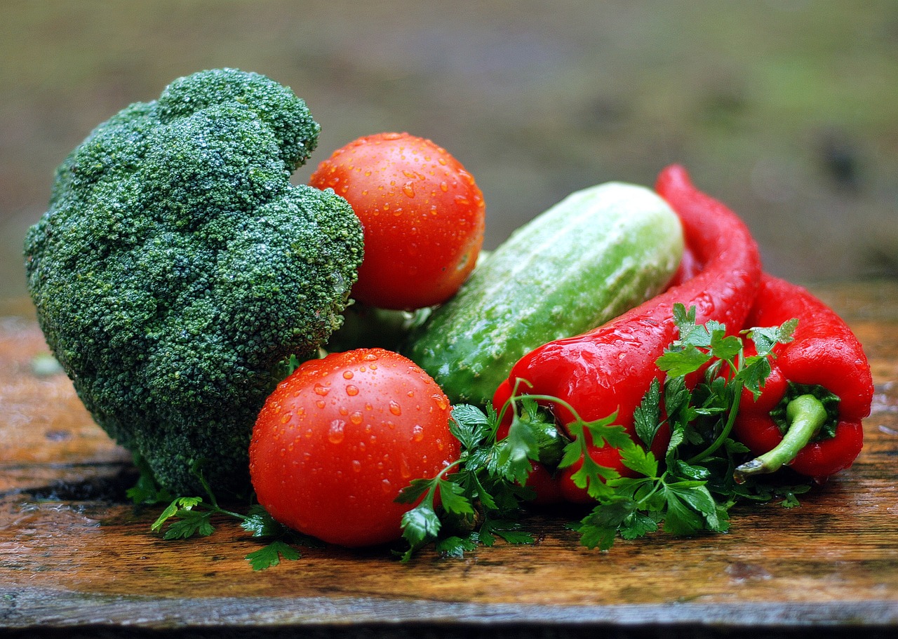 Green Broccoli, Red tomato, Green cucumber, red capsicum and some coriander placed on a brown table in a closed up view. They are wet and very vibrant in colour.